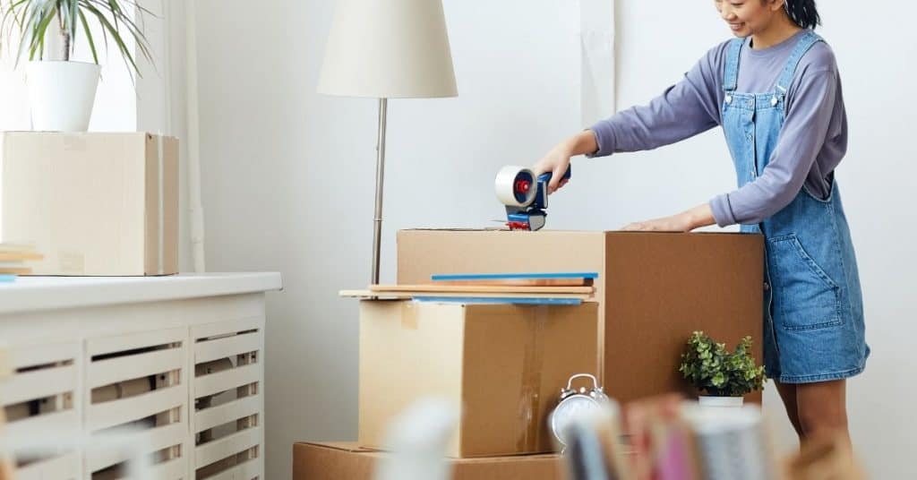 in image you can see a person wearing overalls with a tape roller in their hand closing up a box that is on a table. this is illustrating that the person is cleaning and getting rid of things to declutter their space