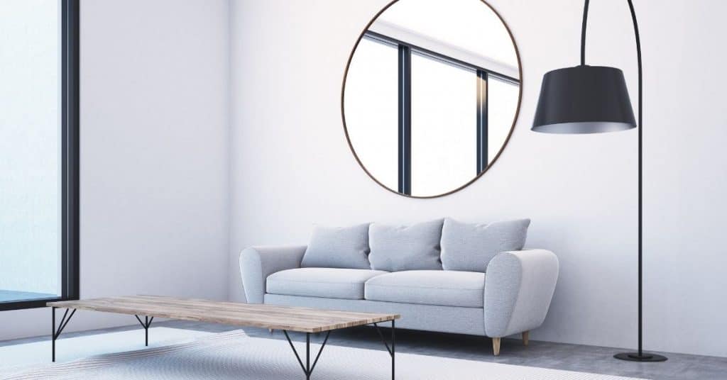 in this image there is a living room that has a sofa and a floor lamp above these items is a mirror that is facing a window. You can see the reflection of the window in the mirror by doing this small tweak a room will look much larger because the mirror will add depth