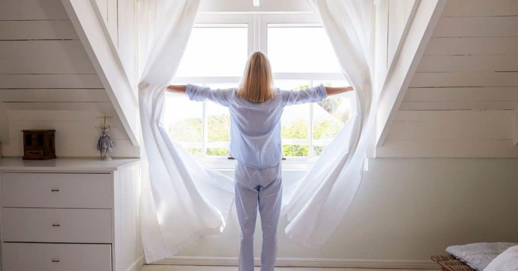 person standing in front of a window that is being opened, curtains are flowing in the room due to breeze coming in from window. Opening windows in a small room can make the room appear larger and bigger than it really is